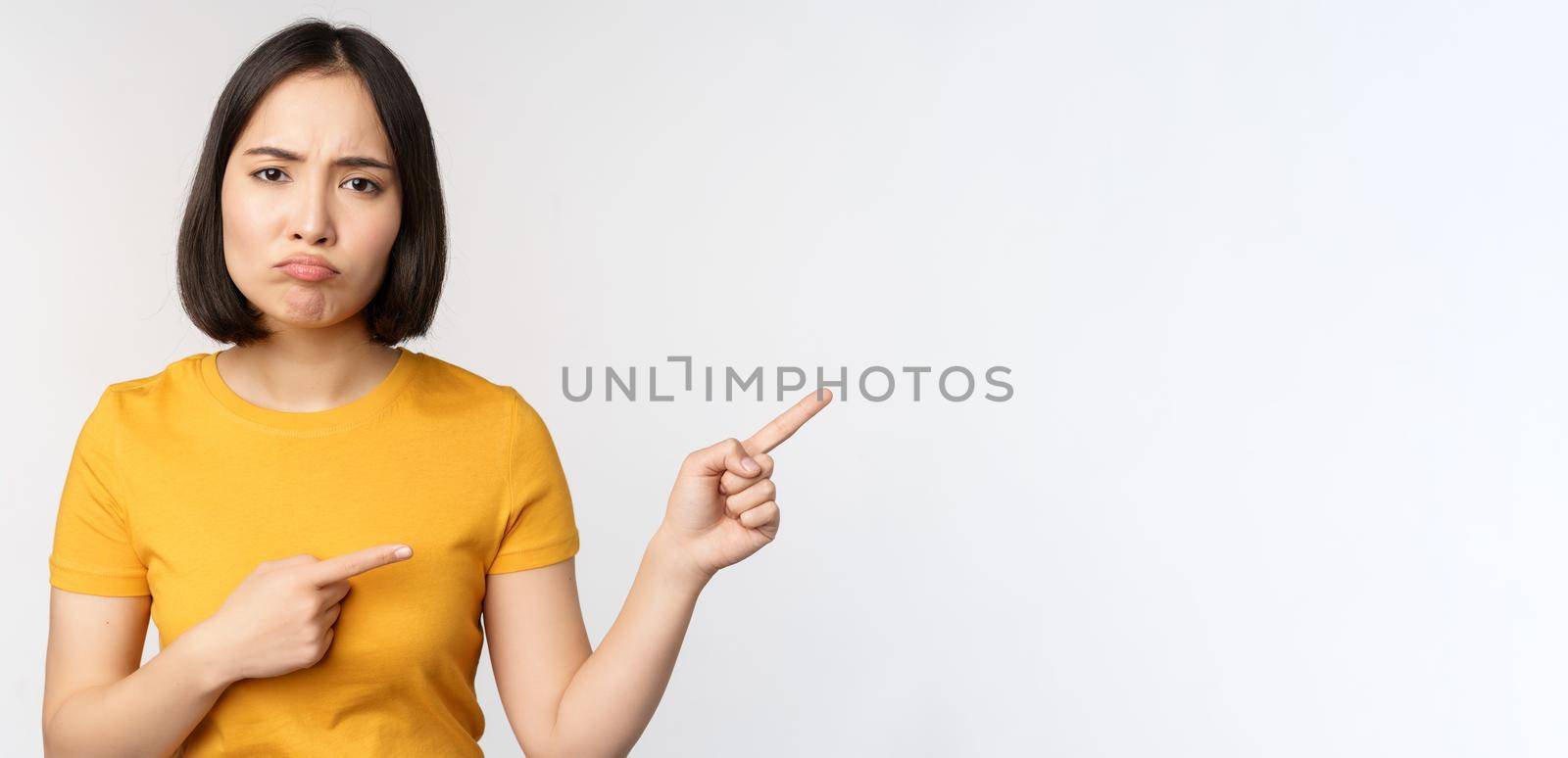 Portrait of disappointed, moody asian girl, pointing fingers right and looking at smth unfair, complaining, standing over white background.