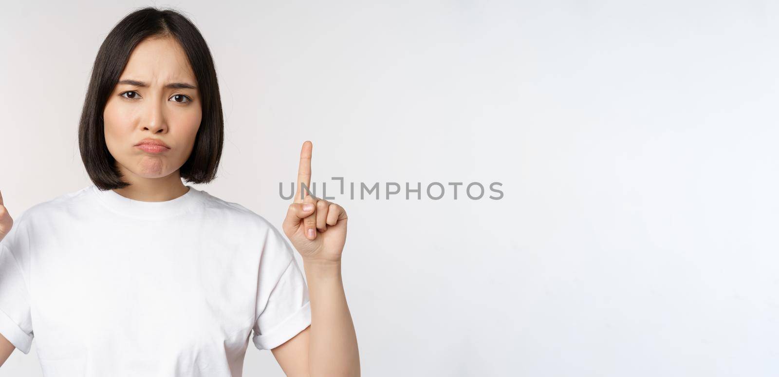 Sad and disappointed young asian woman pointing fingers up, showing advertisement with upset face expression, white studio background by Benzoix