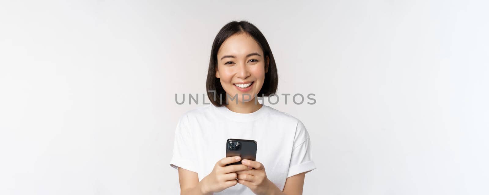 Portrait of smiling asian woman using mobile phone, chatting, texting message, standing in tshirt over white background by Benzoix