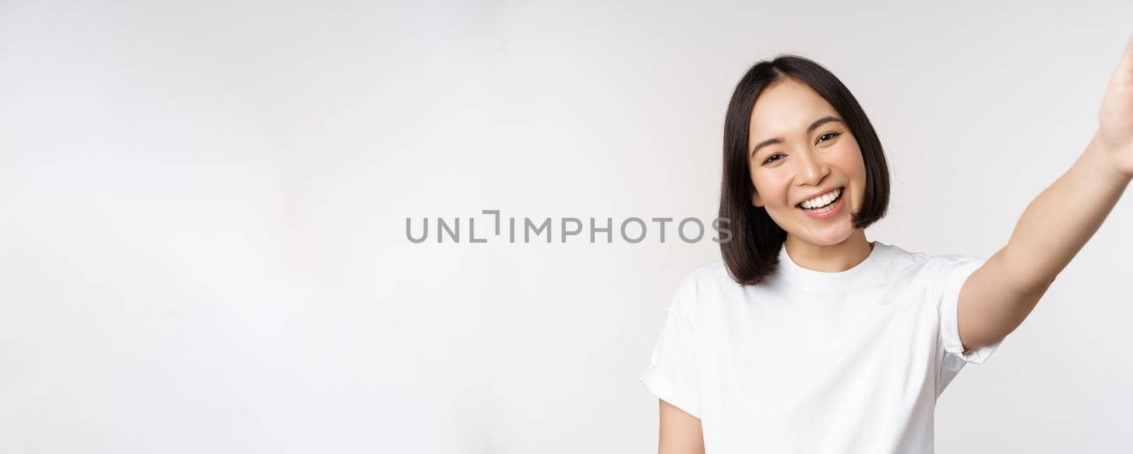 Beautiful young asian woman smiling, looking at camera, holding device, taking selfie, video chat, standing in tshirt over white background by Benzoix