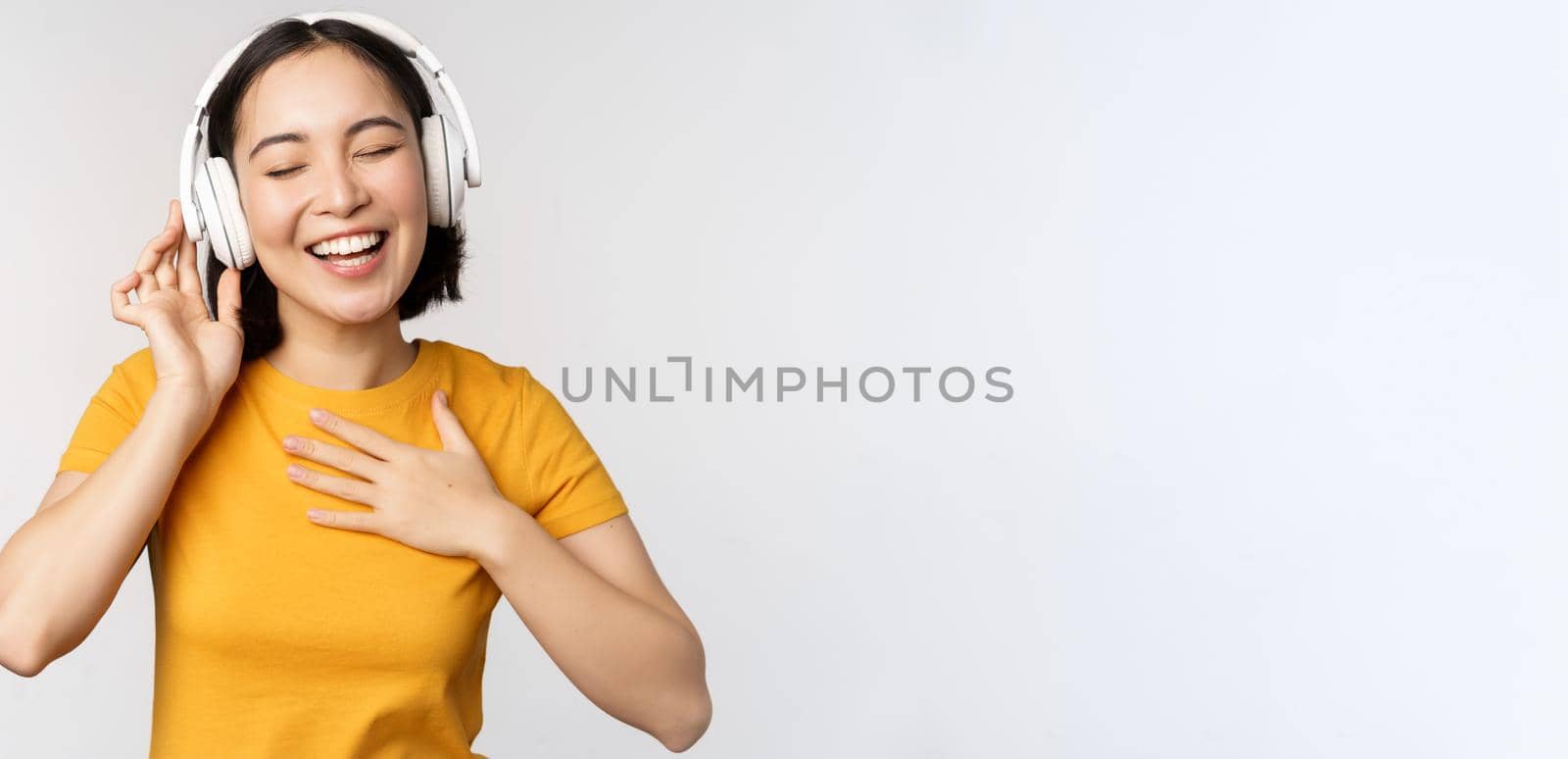 Happy asian girl dancing, listening music on headphones and smiling, standing in yellow tshirt against white background by Benzoix