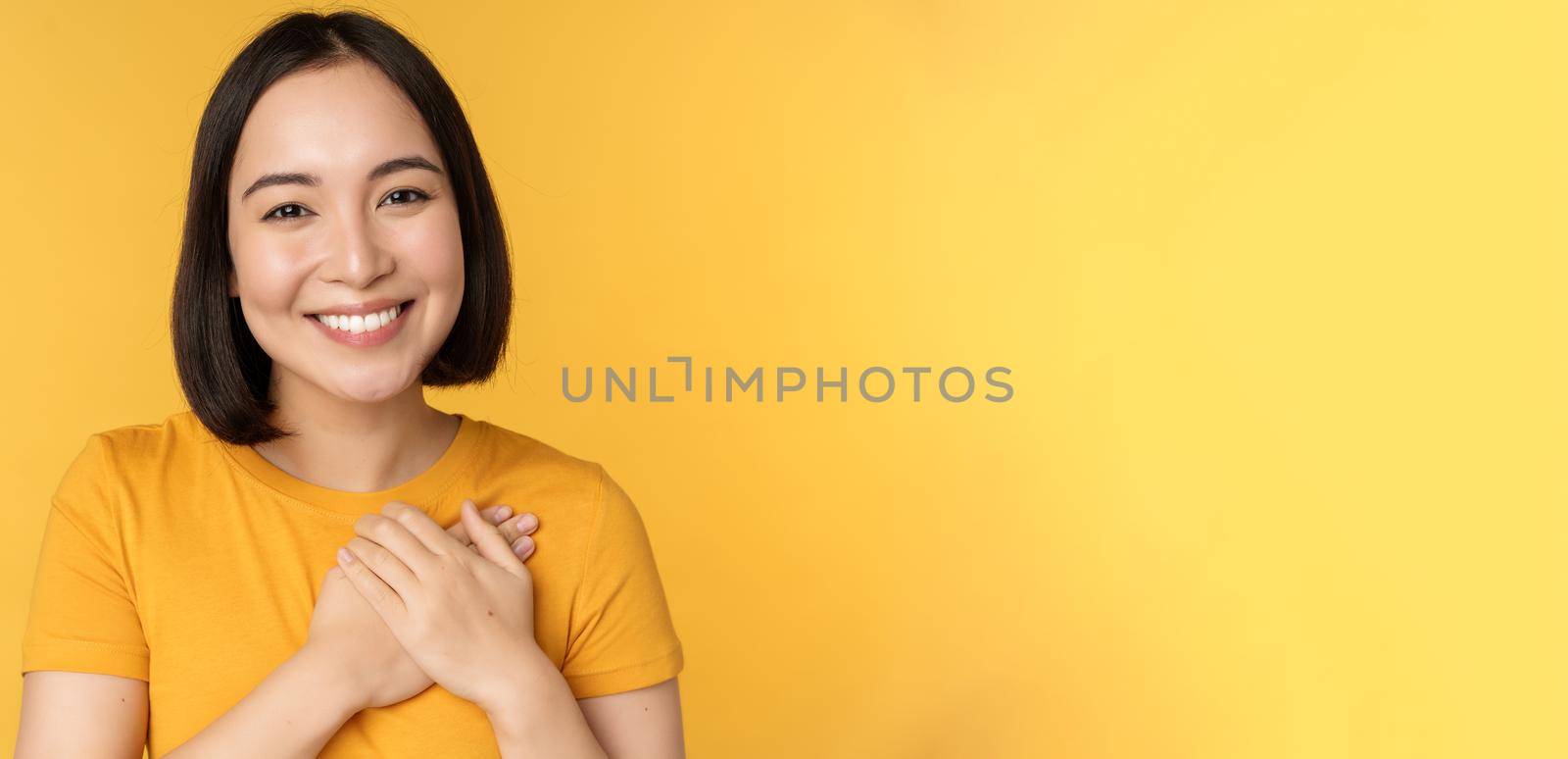 Beautiful asian woman, smiling with tenderness and care, holding hands on heart, standing in tshirt over yellow background by Benzoix