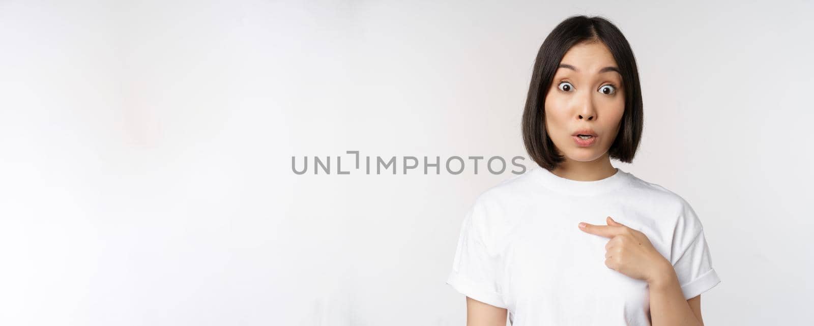 Portrait of surprised asian girl student, pointing finger left, raising eyebrows amazed, impressed by big deal, offer ahead, standing over white background.