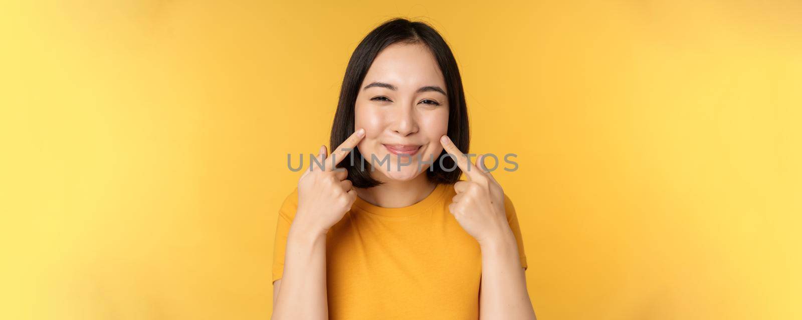 Close up portrait of cute asian girl showing her dimples and smiling coquettish at camera, standing over yellow background by Benzoix