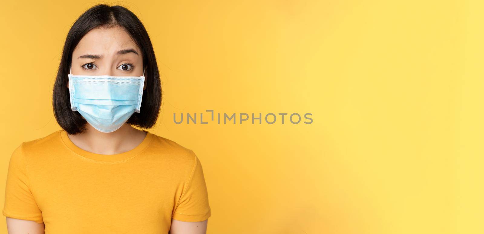 Portrait of skeptical and confused asian woman in medical face mask, raising eyebrow doubtful, standing over yellow background.