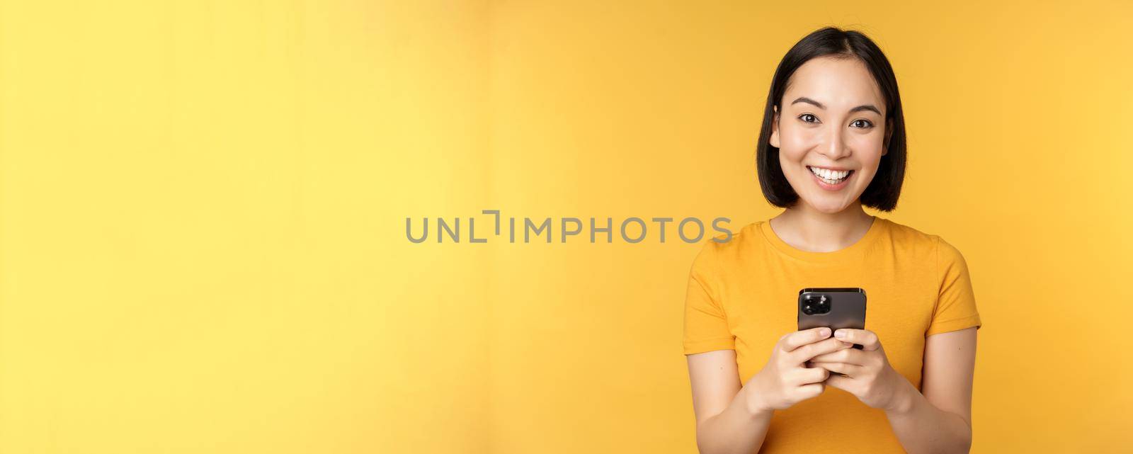 Technology. Smiling asian woman using mobile phone, holding smartphone in hands, standing in t-shirt against yellow background by Benzoix