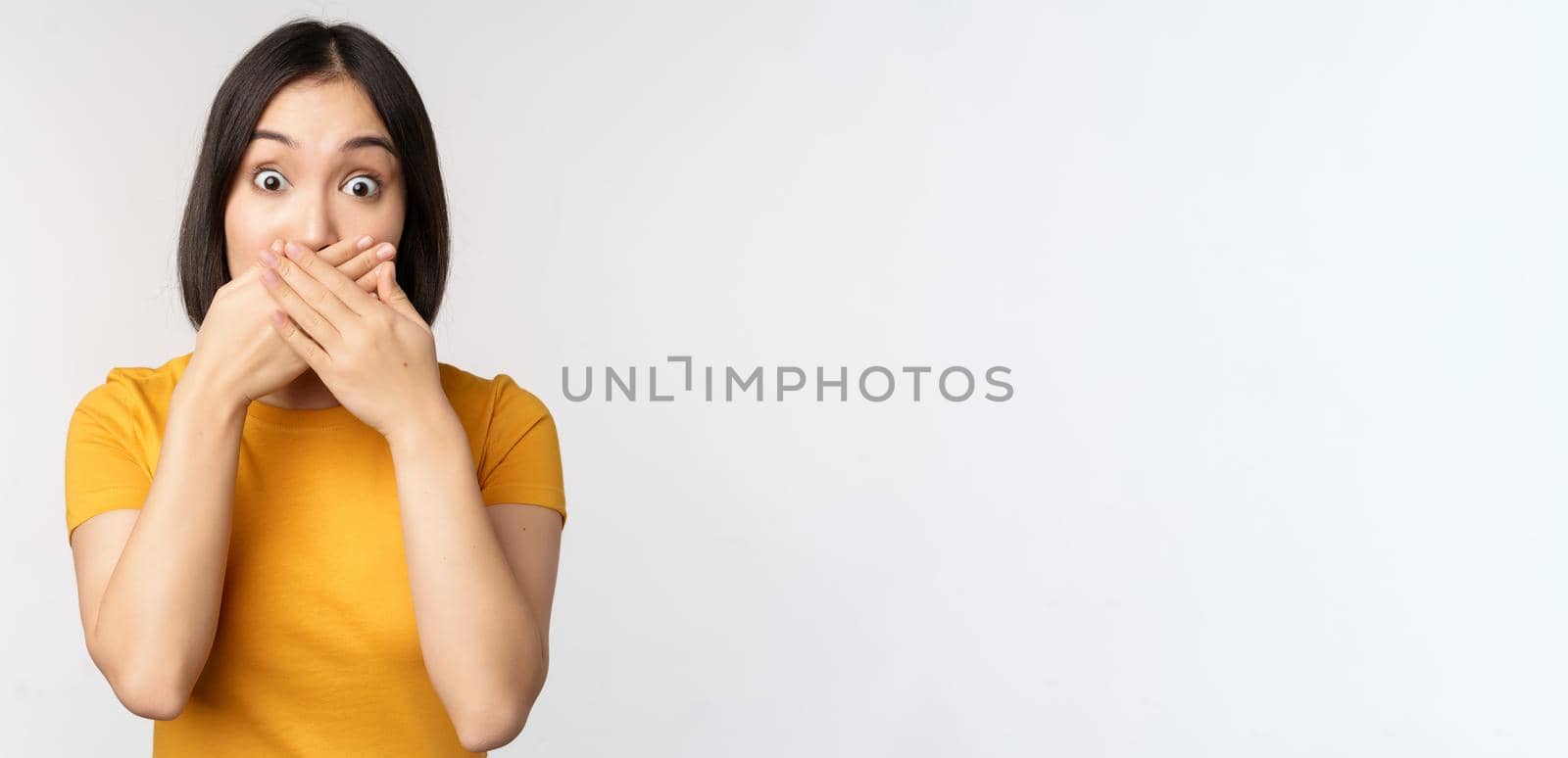 Shocked asian woman cover mouth with hands, looking startled with speechless face expression, standing in yellow tshirt against white background.