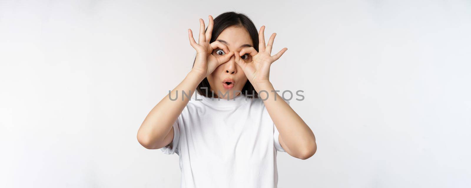 Funny young asian woman, korean girl making eyes glasses gesture, looking happy at camera, standing over white background.