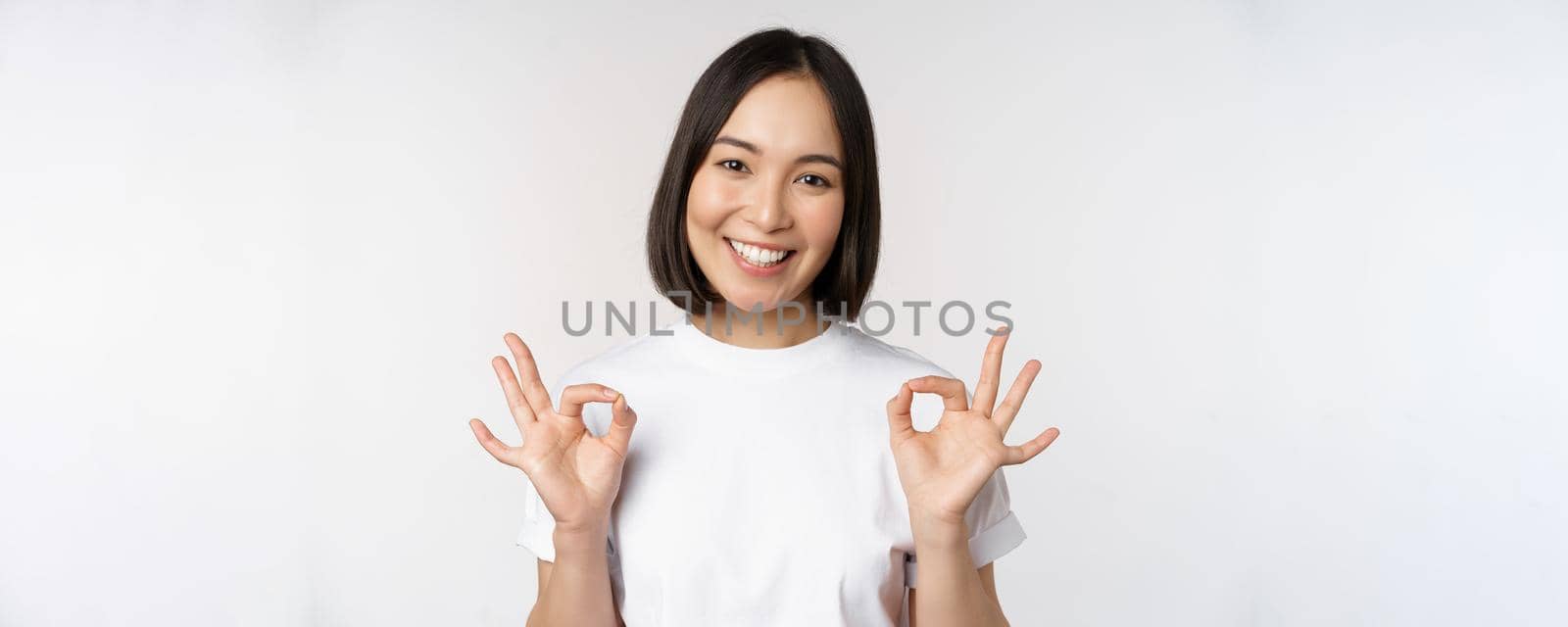 Very well, excellent. Smiling asian woman showing okay sign, approval, ok gesture, looking satisfied, recommending smth, standing over white background by Benzoix