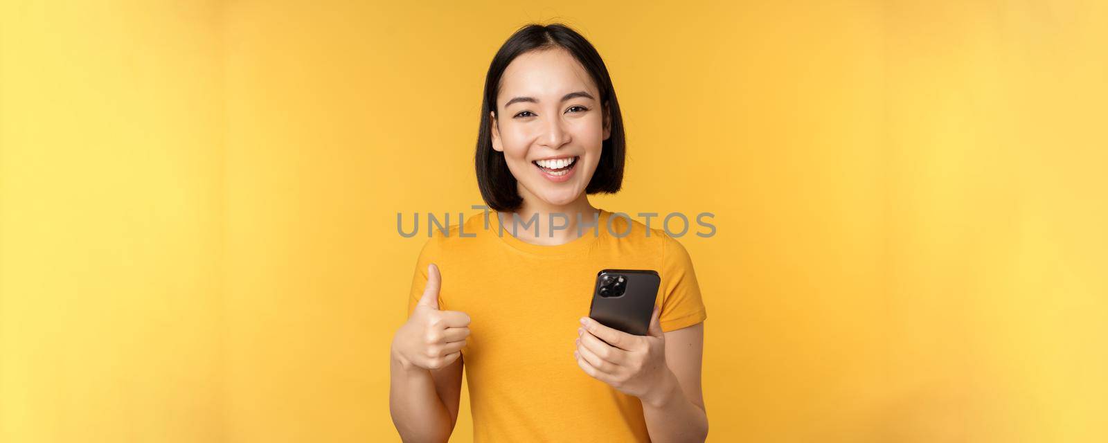 Happy smiling asian girl holding mobile phone and showing thumbs up, recommending application on smartphone, standing over yellow background.