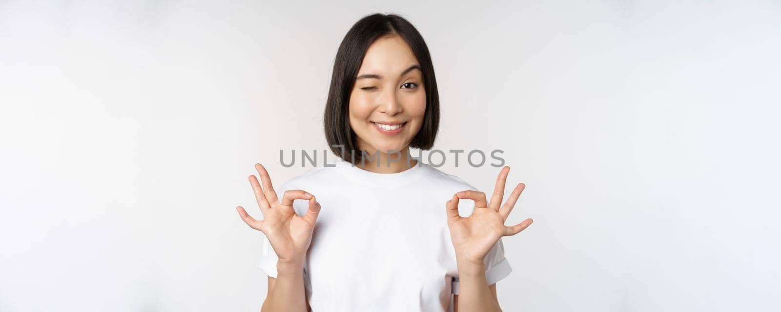 Very well, excellent. Smiling asian woman showing okay sign, approval, ok gesture, looking satisfied, recommending smth, standing over white background by Benzoix