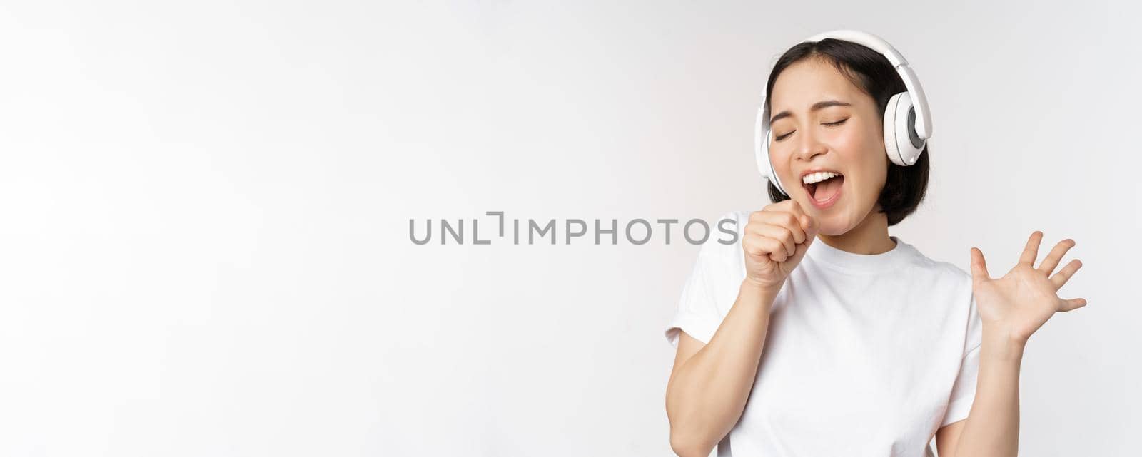 Korean girl sings and listents music in headphones, having fun, stands over white background. Copy space
