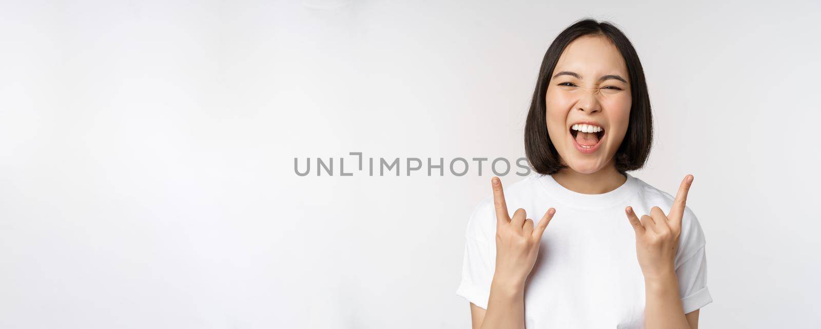 Sassy asian girl shouting, enjoying concert or festival, showing rock on, heavy metal sign, having fun, standing over white background by Benzoix