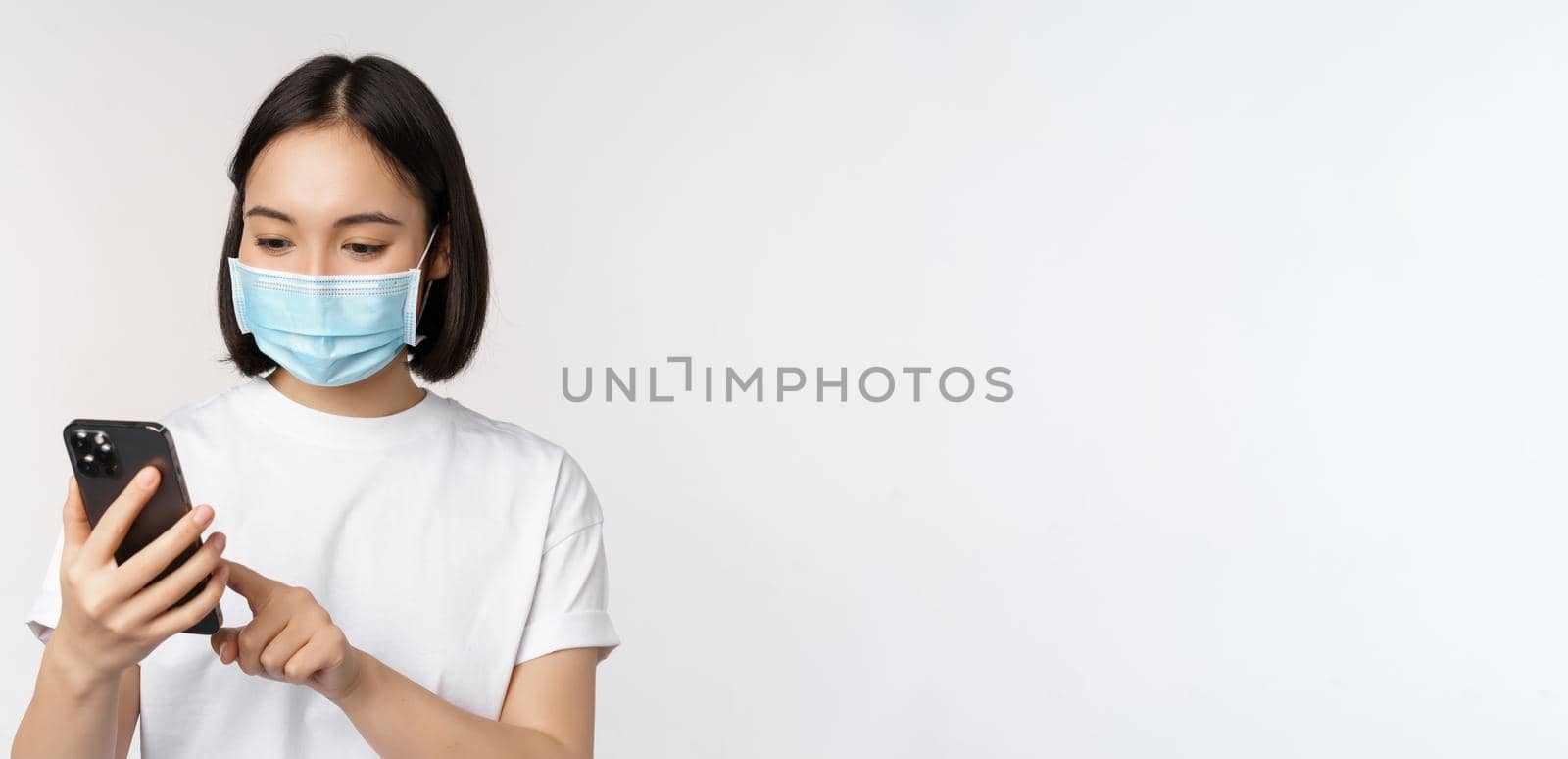 Health and covid-19 concept. Young asian woman in medical mask using mobile phone, typing on smartphone, standing against white background.