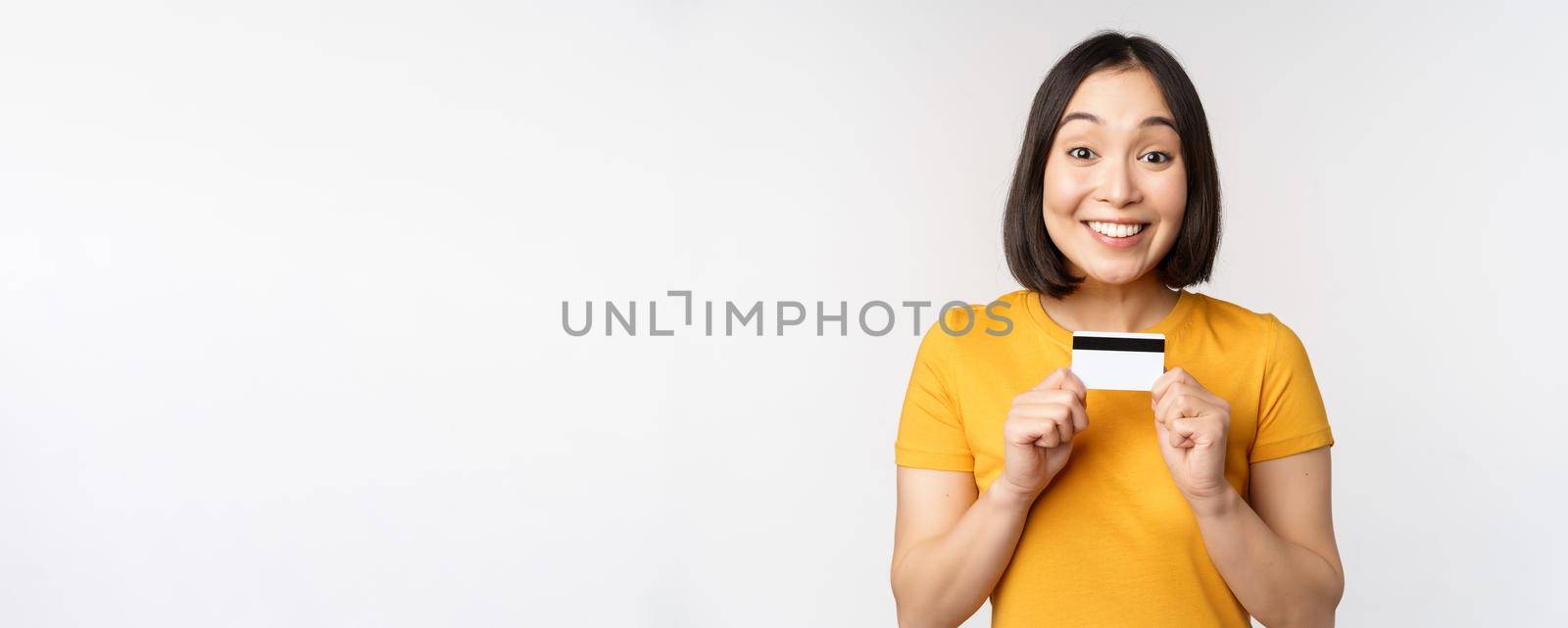 Portrait of beautiful korean girl holding credit card, recommending bank service, standing in yellow tshirt over white background.