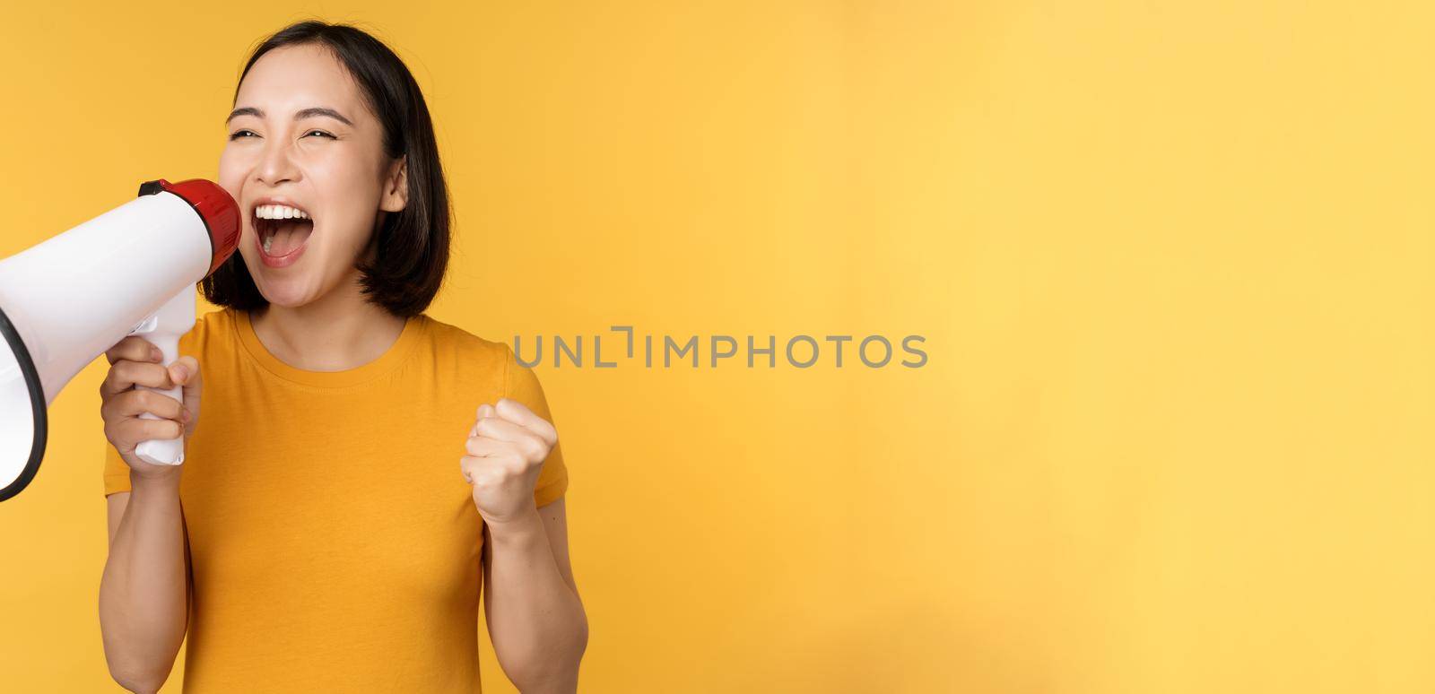 Announcement. Happy asian woman shouting loud at megaphone, recruiting, protesting with speaker in hands, standing over yellow background.