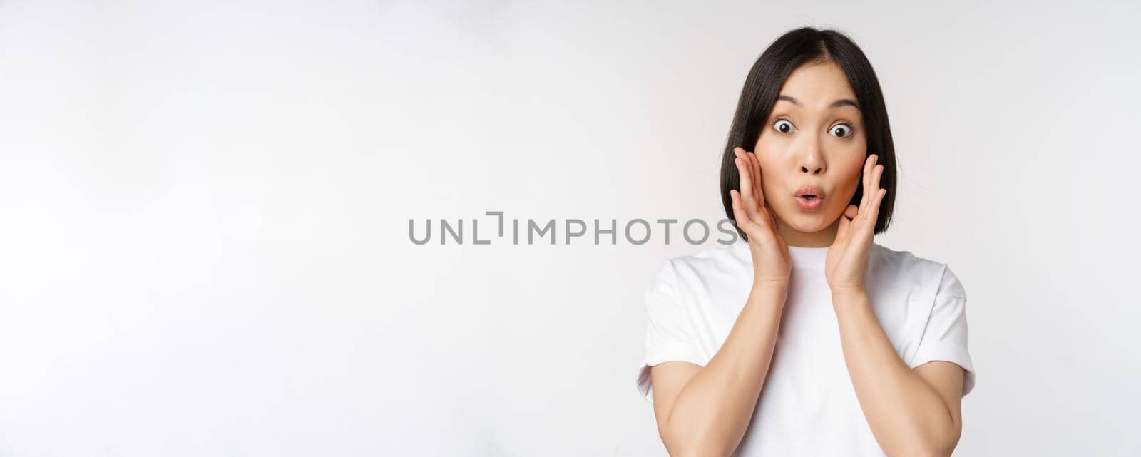 Close up portrait of asian girl looking surprised, wow face, reacting amazed at smth, standing in white tshirt over studio background, isolated.
