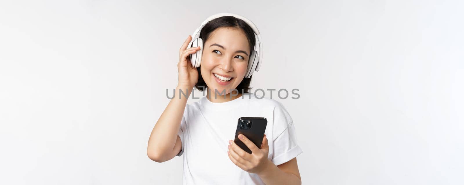 Beautiful modern asian girl, listening music in headphones, holding mobile phone, using smartphone app, standing over white background.