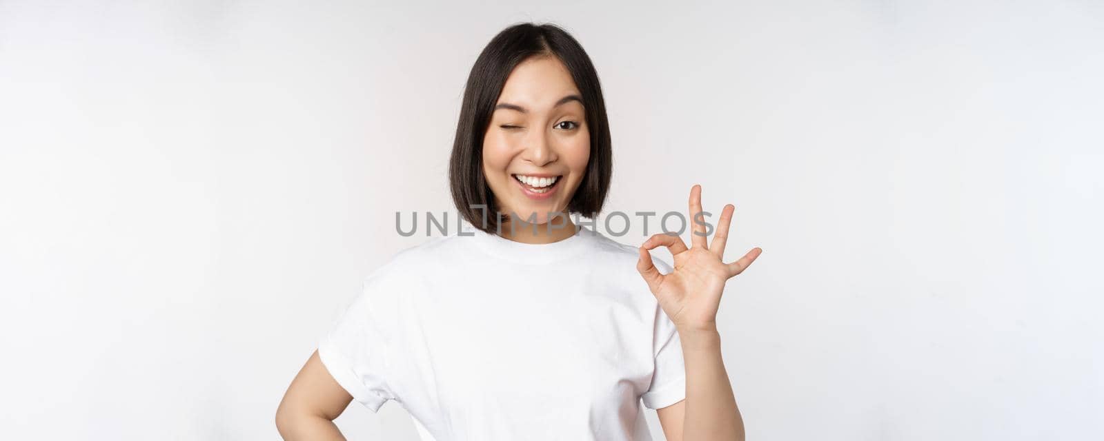 Everything okay. Smiling young asian woman assuring, showing ok sign with satisfied face, standing over white background by Benzoix