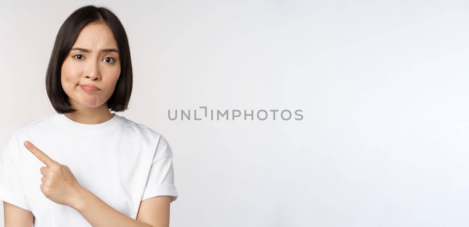 Skeptical asian girl in white t-shirt, pointing at product or logo with disappointed grimace, dislike and complain at smth, standing over white background by Benzoix