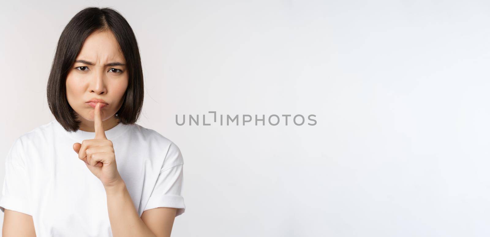 Angry asian girl shushing, keep quiet, taboo silence gesture, press finger to lips and frowning, scolding for being too loud, standing over white background.