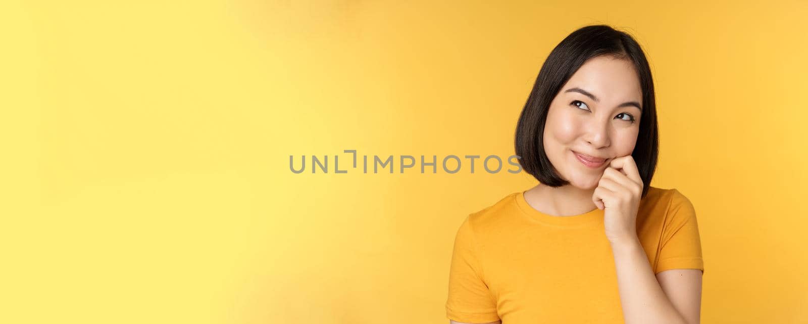 Close up portrait of cute asian girl smiling, thinking, looking up thoughtful, standing in tshirt over yellow background. Copy space