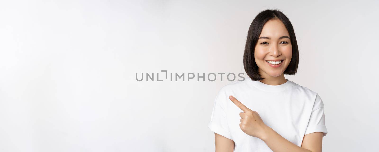 Close up of young japanese woman smiling, pointing finger left at copy space, showing announcement or advertisment banner, standing over white background.