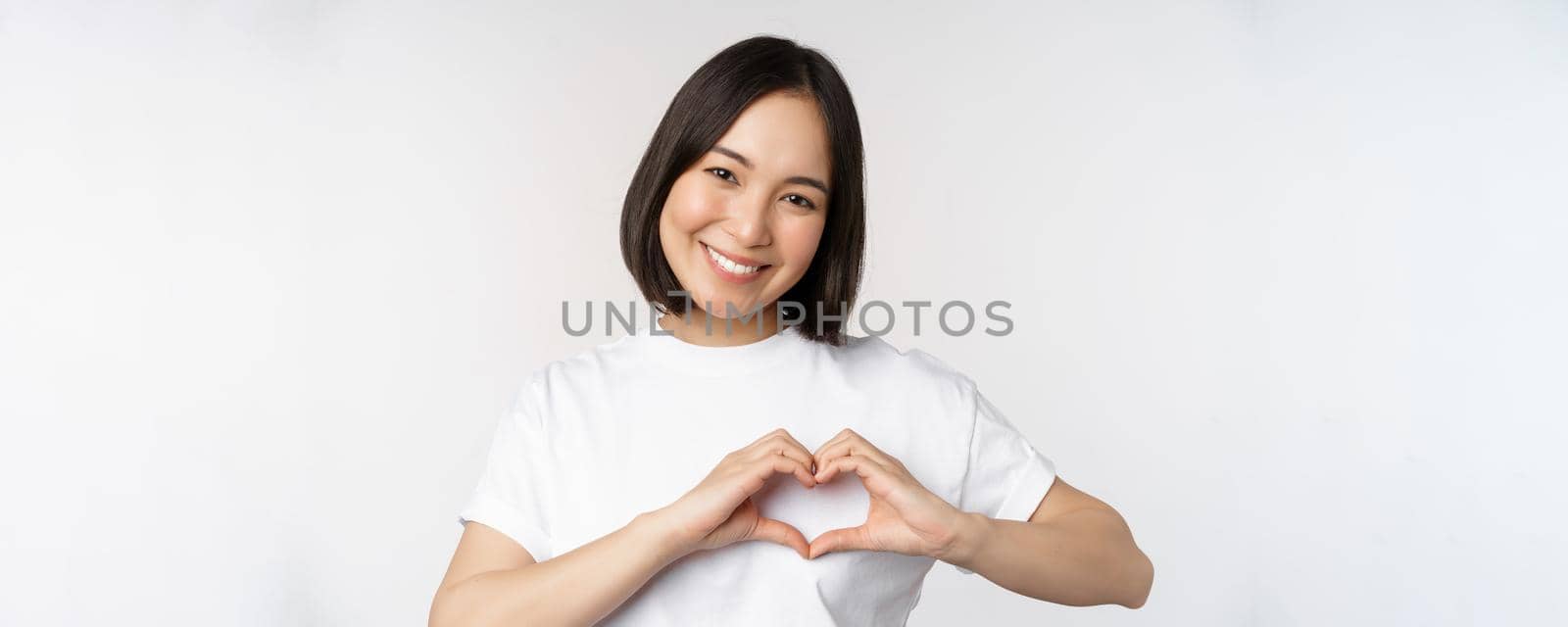 Lovely asian woman smiling, showing heart sign, express tenderness and affection, standing over white background.
