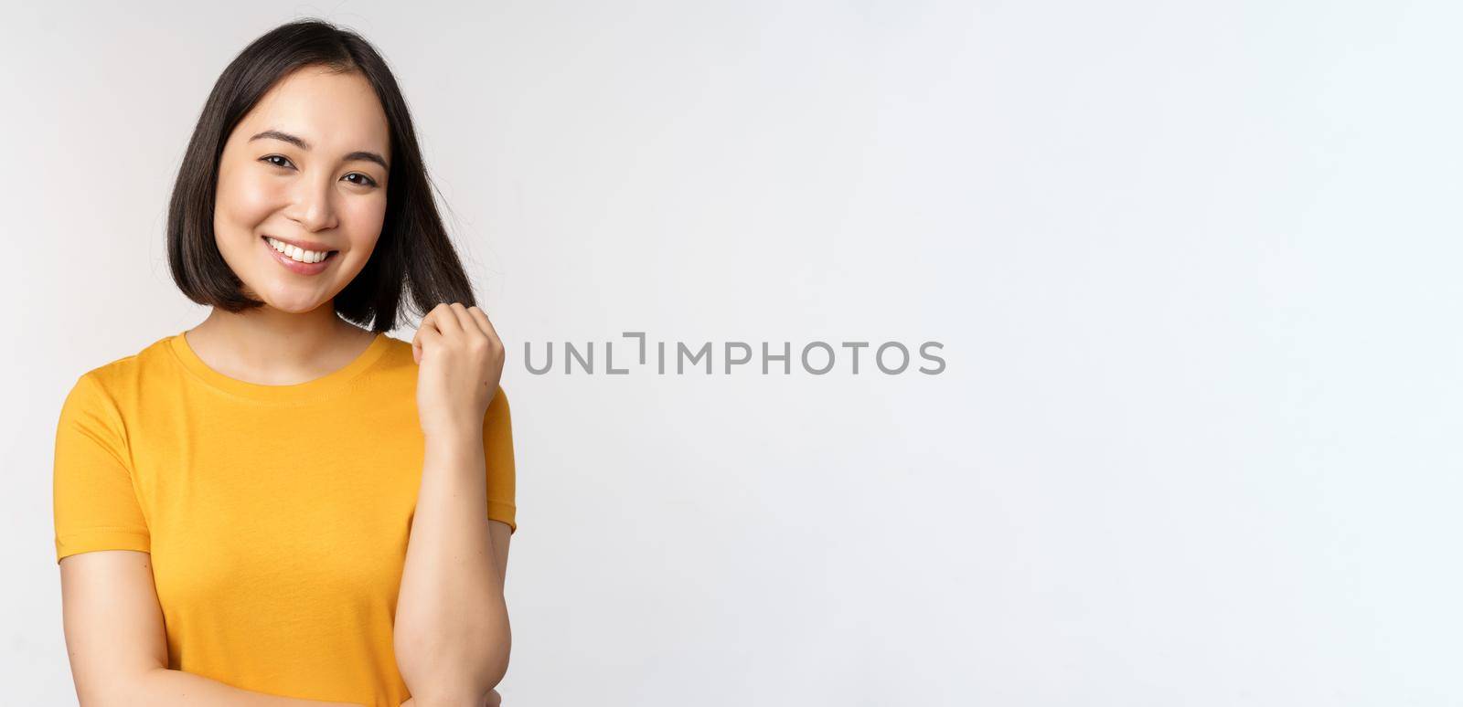 Beautiful romantic asian girl, smiling and playing with hair, looking happy at camera, standing in yellow t-shirt over white background by Benzoix