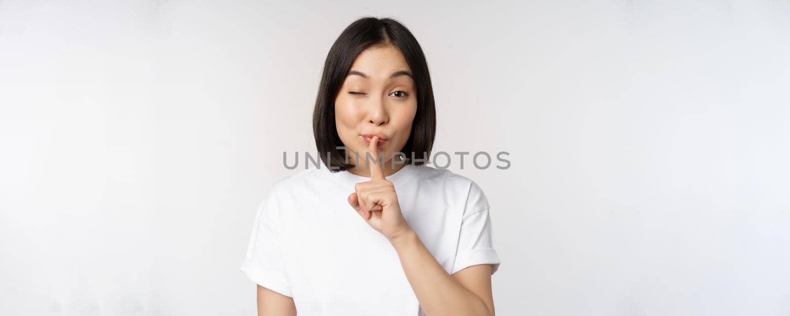 Close up portrait of young beautiful asian girl shushing, has secret, keep quiet silence gesture, press finger to lips, standing in tshirt over white background.