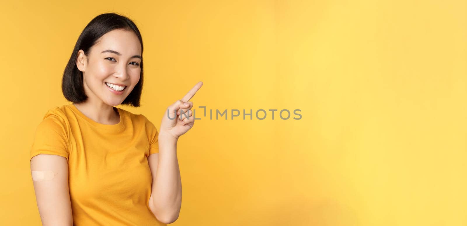 Vaccination and covid-19 pandemic concept. Smiling korean woman with band aid on shoulder after coronavirus vaccine shot, pointing at banner with vaccinating campaign.