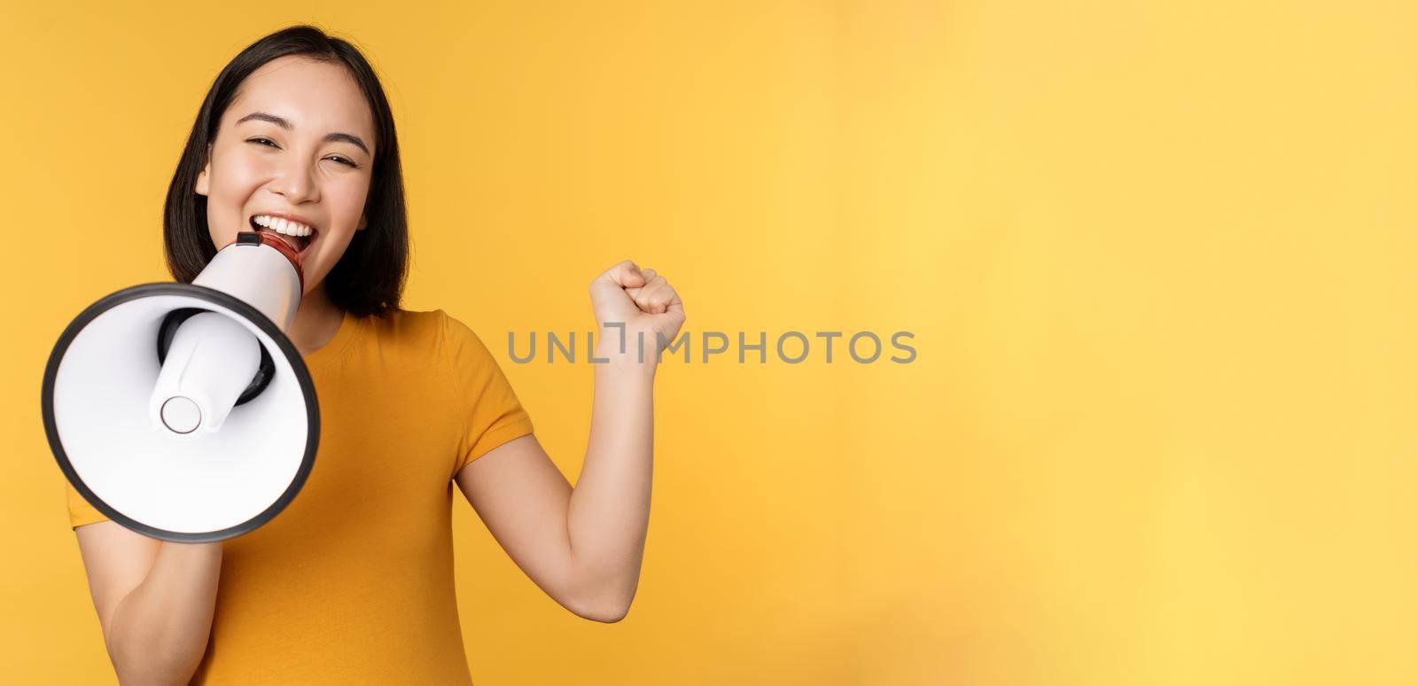 Smiling asian woman standing with megaphone, announcing smth, advertising product, standing over yellow background by Benzoix