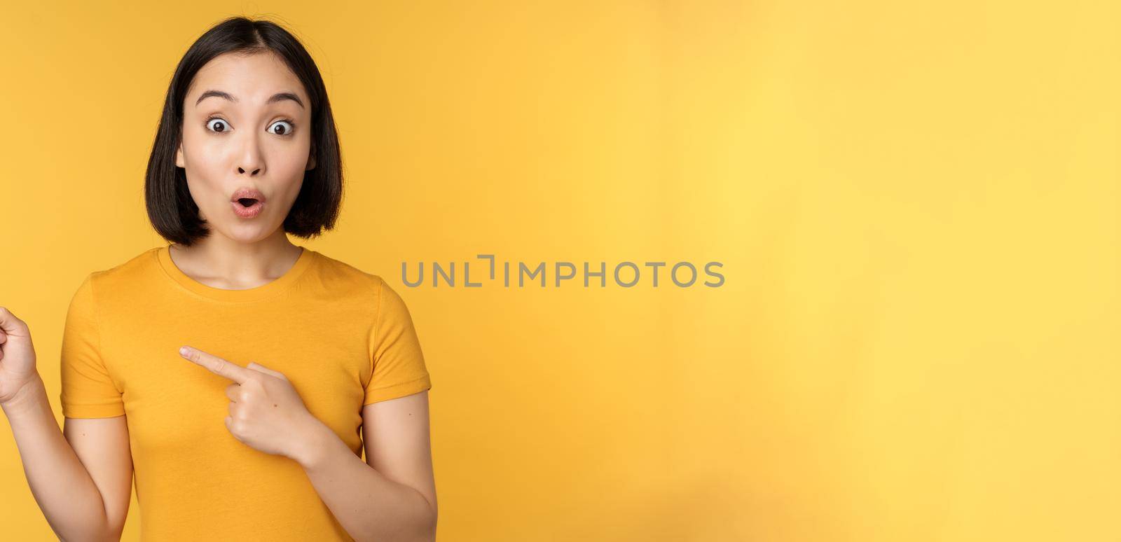 Smiling asian woman pointing fingers left, showing advertisement on empty copy space, standing over yellow background.