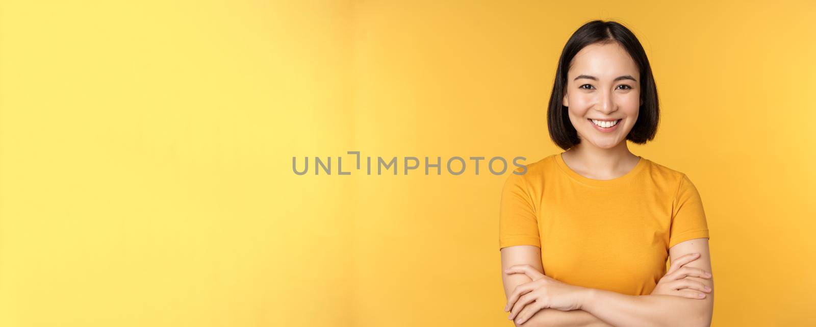 Confident asian girl cross arms on chest, smiling and looking assertive, standing over yellow background by Benzoix