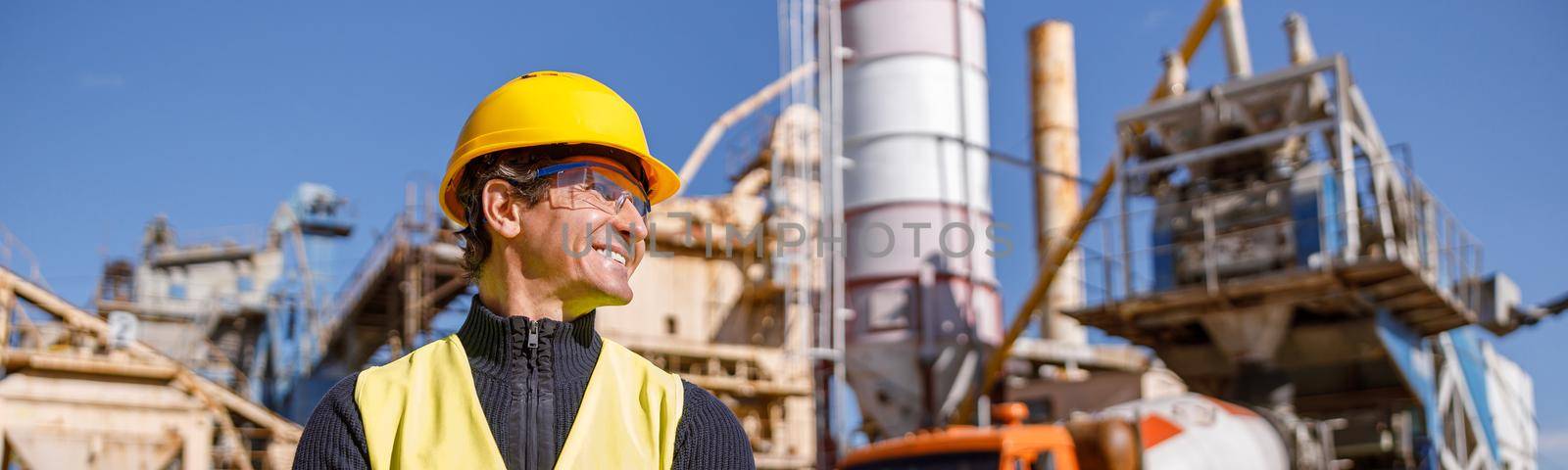 Joyful matured man factory worker wearing safety helmet and work vest while keeping arms crossed and smiling