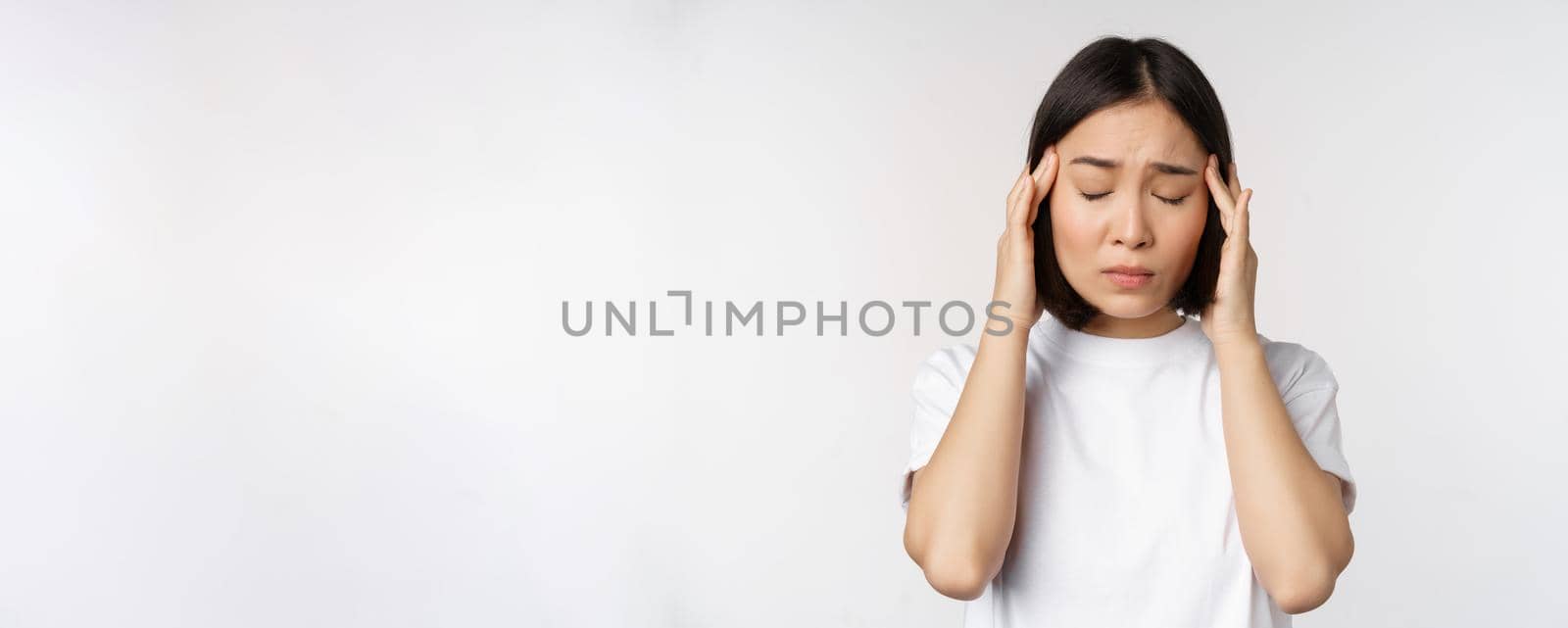 Portrait of asian girl feeling headache, migraine or being ill, standing in white t-shirt over white background. Copy space