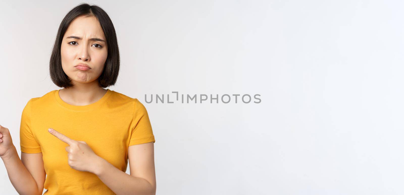 Portrait of disappointed brunette asian girl, pointing fingers left, grimacing upset, showing smth unpleasant, standing over white background by Benzoix