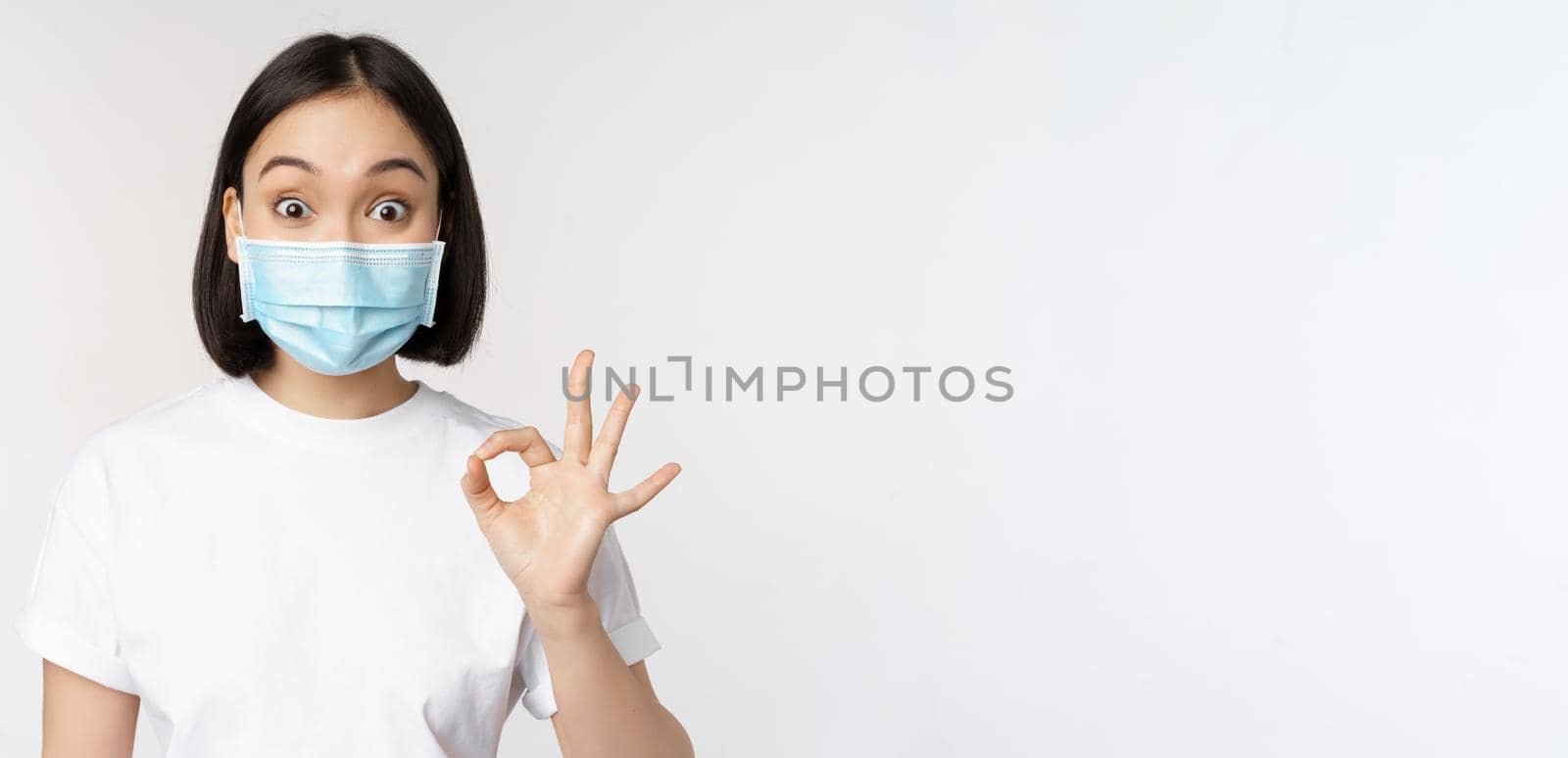 Covid-19, healthcare and medical concept. Impressed asian woman in medical mask, looking amazed and showing ok, okay sign, standing over white background by Benzoix