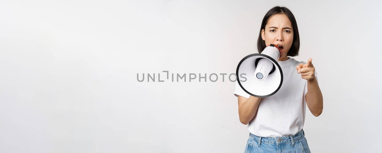 Angry asian woman with megaphone, scolding, accusing someone, protesting with speakerphone on protest, standing over white background.