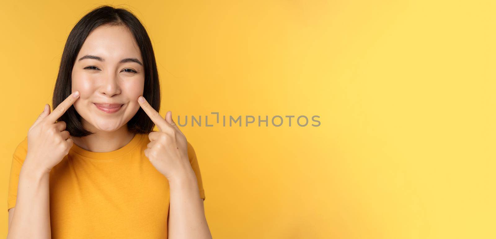 Close up portrait of cute asian girl showing her dimples and smiling coquettish at camera, standing over yellow background. Copy space