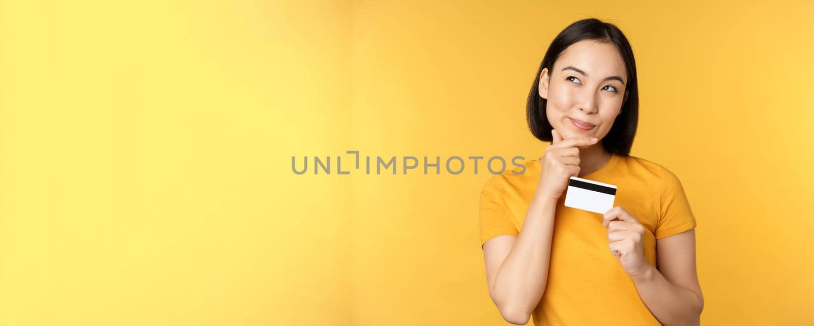 Thinking asian woman holding credit card and smiling, planning buy smth, standing thoughtful over yellow background.
