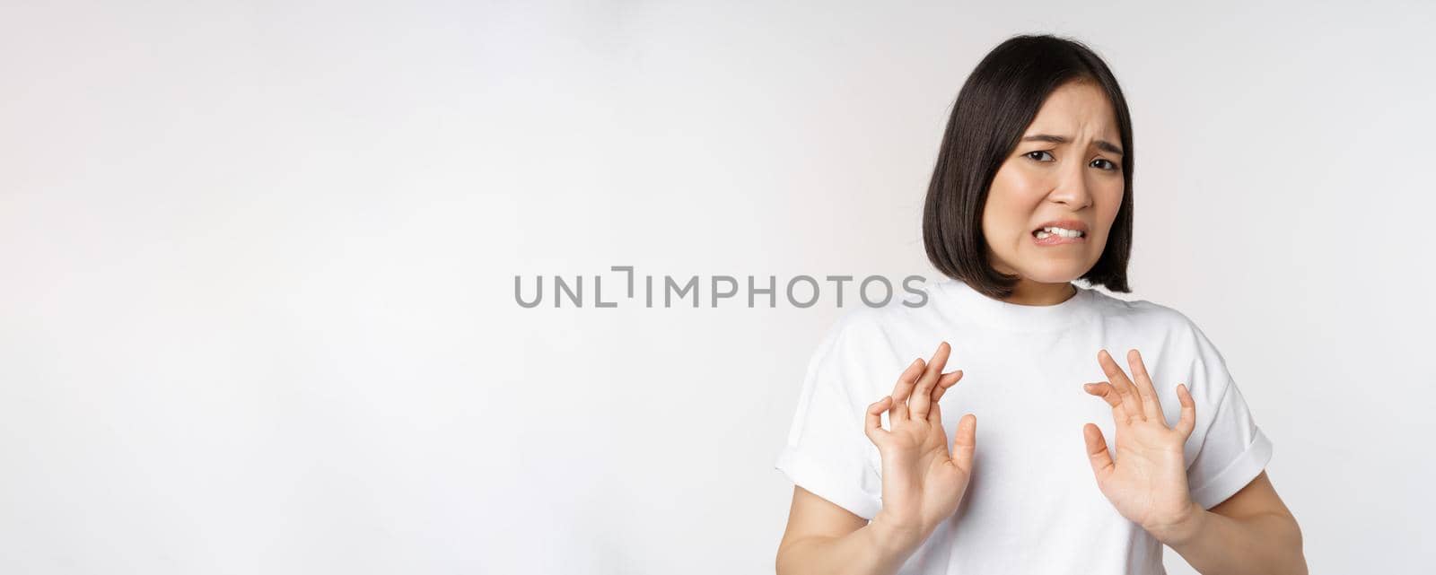 Disgusted asian woman rejecting smth, grimacing from dislike and aversion, stare with cringe, refusing offer, standing over white background.