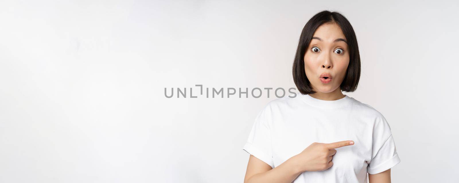 Portrait of surprised asian woman say wow, pointing right at amazing product offer, sale or logo, standing over white background.