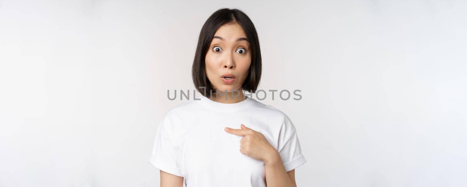 Portrait of surprised asian girl student, pointing finger left, raising eyebrows amazed, impressed by big deal, offer ahead, standing over white background by Benzoix