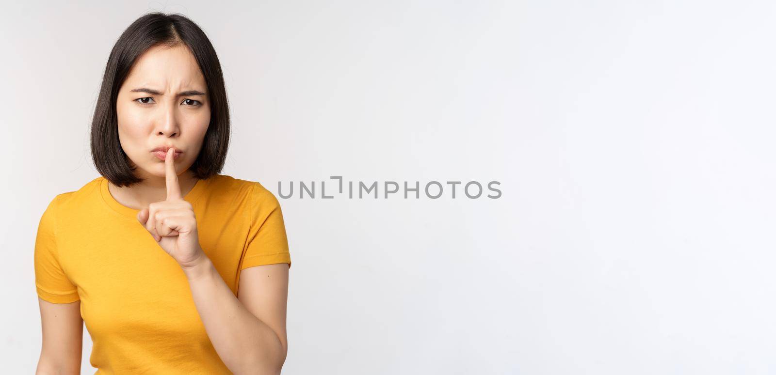 Portrait of angry asian woman shushing, press finger to lips, taboo stop talking sig, looking annoyed, scolding, standing over white background.