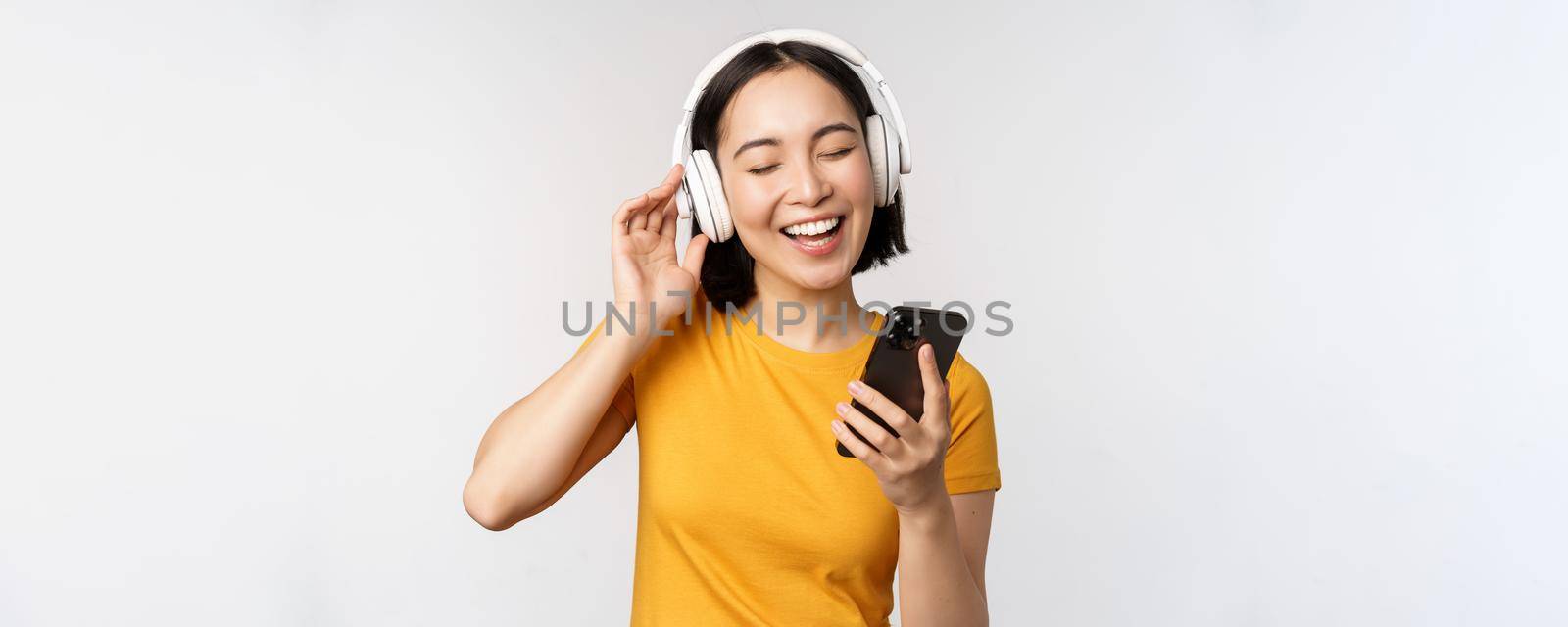 Cute japanese girl in headphones, looking at mobile phone and smiling, using music app on smartphone, standing against white background.