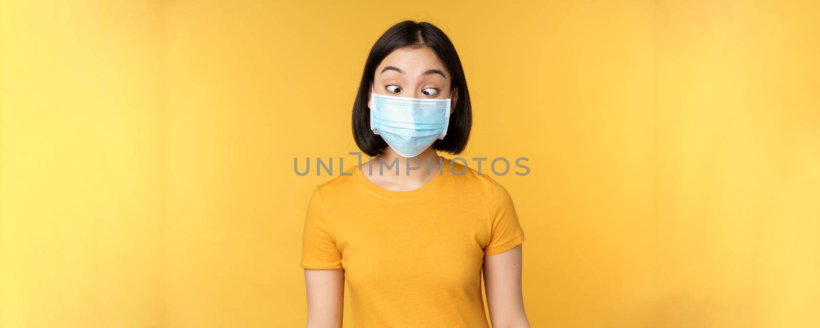 Health and people concept. Funny asian girl squinting, looking at her face medical mask, standing over yellow background.