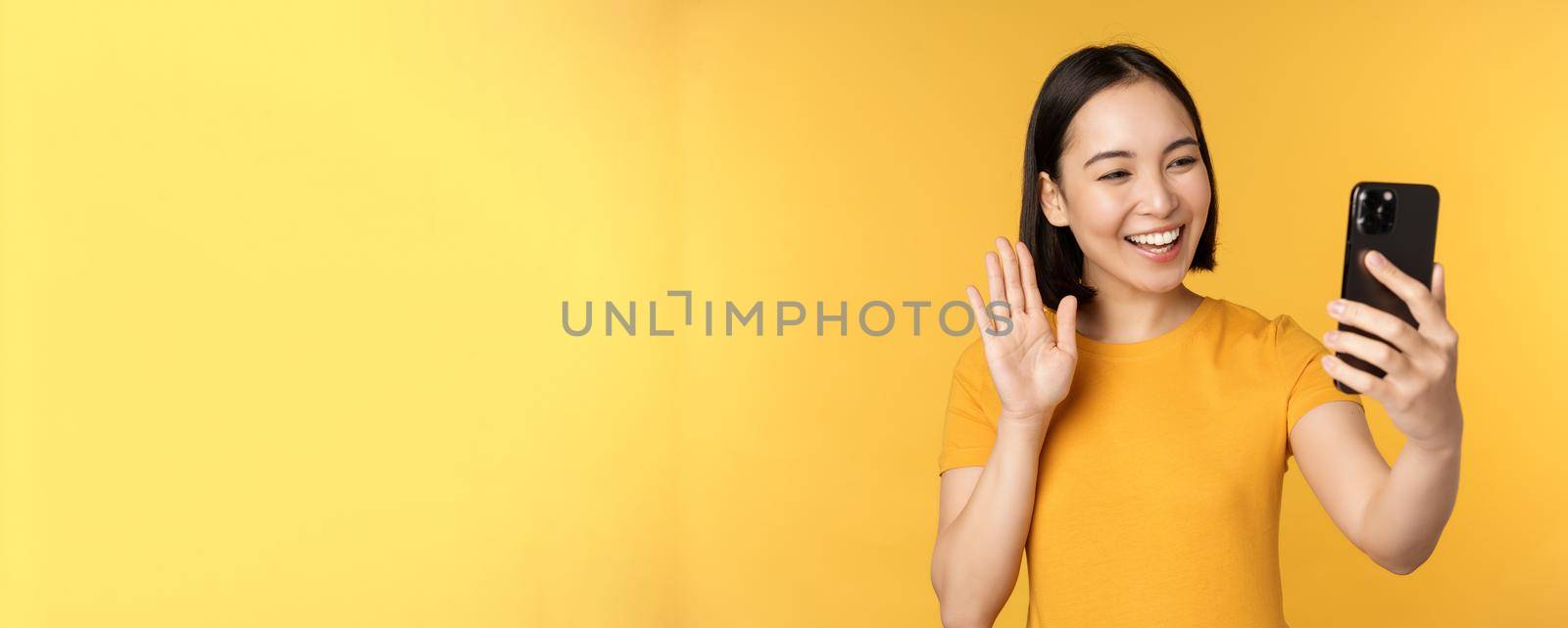 Image of happy, beautiful asian girl video chatting, talking on smartphone application, standing against yellow background. Copy space