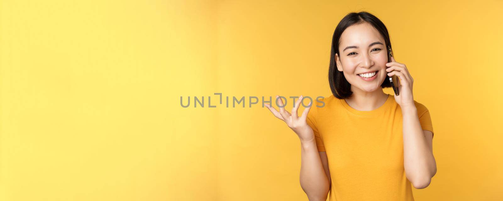 Young korean girl talking on mobile phone. Asian woman calling on smartphone, standing over yellow background by Benzoix