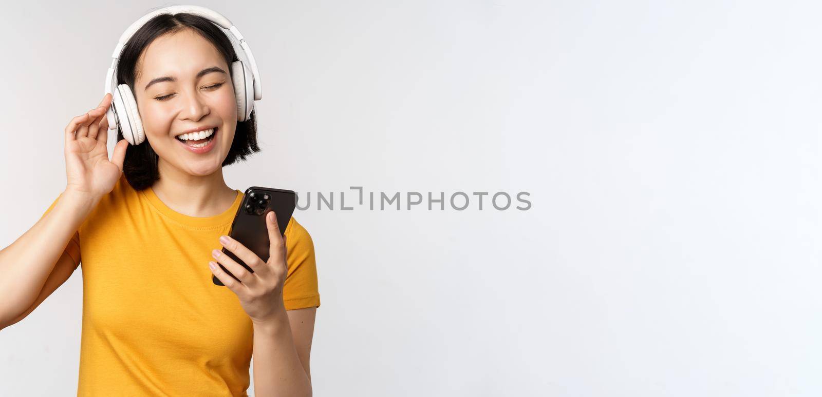 Cute japanese girl in headphones, looking at mobile phone and smiling, using music app on smartphone, standing against white background by Benzoix